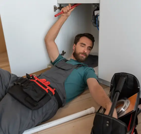 Plumber lying on the floor, reaching for tools in a tool bag while working under a sink cabinet.